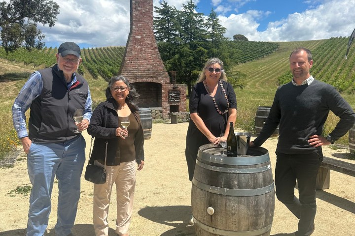 Four people by a wine barrel outdoors, vineyard background with brick chimney.