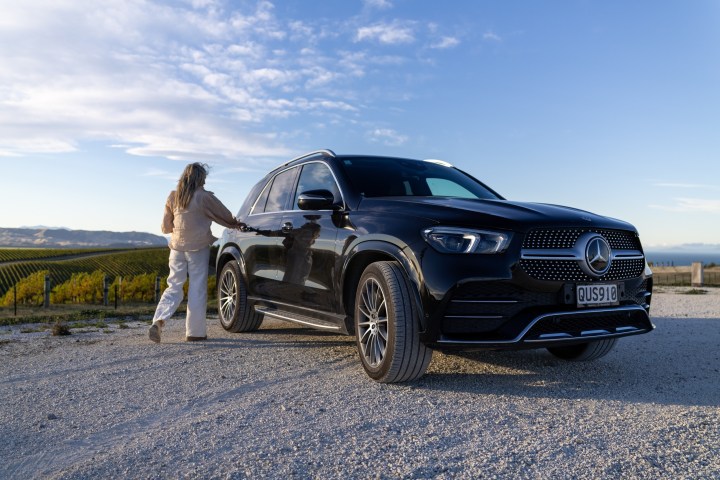 Person near black Mercedes SUV on gravel road with vineyard background.