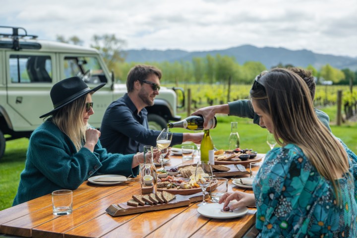 People enjoying a meal outdoors at a wooden table near a vineyard with mountains in the background.