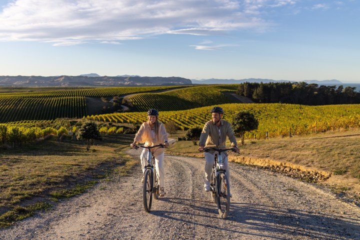 Two people biking on a dirt path with vineyards and hills in the background.